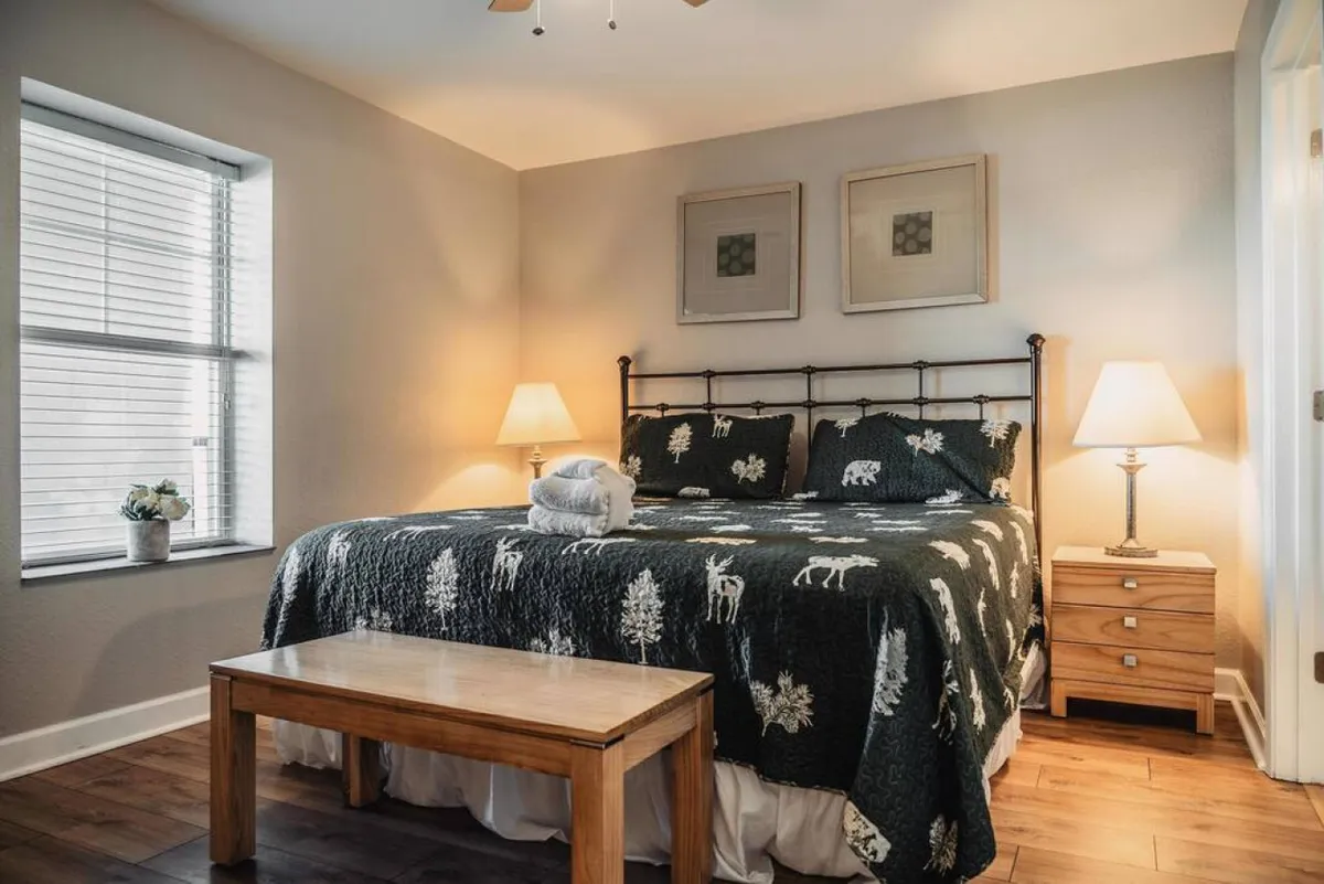 The image showcases a bright and inviting bedroom featuring a plush bed adorned with patterned bedding, a cozy wooden bedside table, and a softly glowing lamp. Sunlight filters through, enhancing the warmth and comfort of the space, inviting guests to relax, read, or savor a morning coffee in bed.