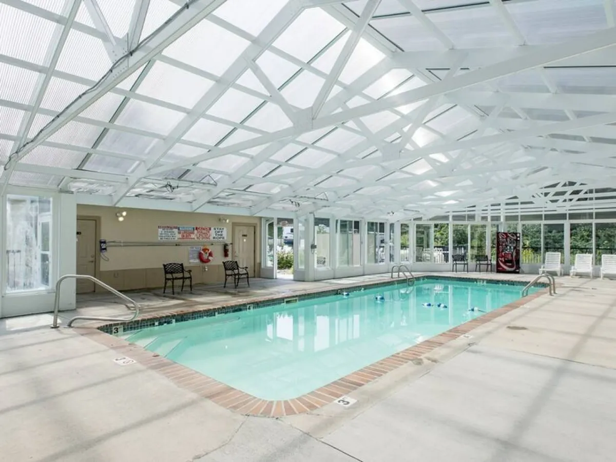 A spacious, heated indoor swimming pool with a clear roof structure, allowing plenty of natural light to flood the area. Comfortable lounge chairs line the pool's edge, and a lifeguard station is visible, creating a safe and family-friendly atmosphere.