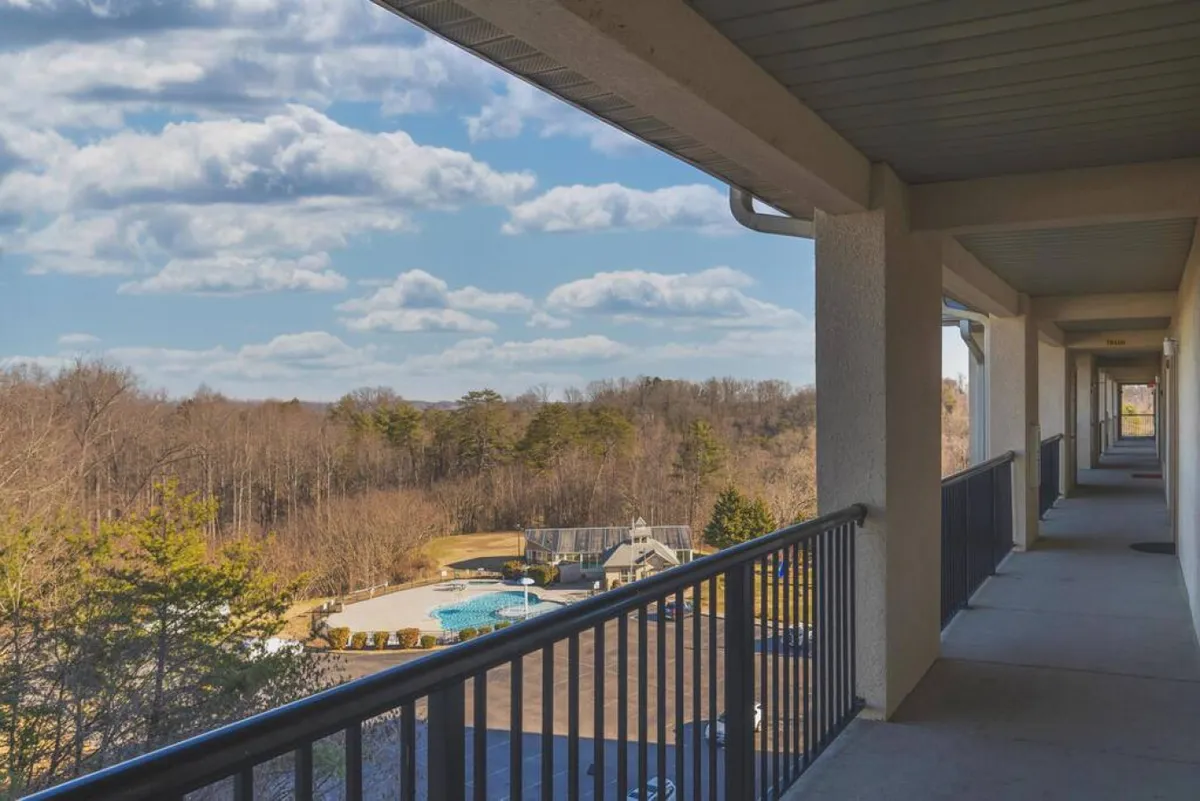 A view from a private balcony with a railing, overlooking a sparkling swimming pool, surrounded by lush forest and an expansive sky with clouds. The scene suggests a peaceful retreat with access to both scenic vistas and outdoor amenities.