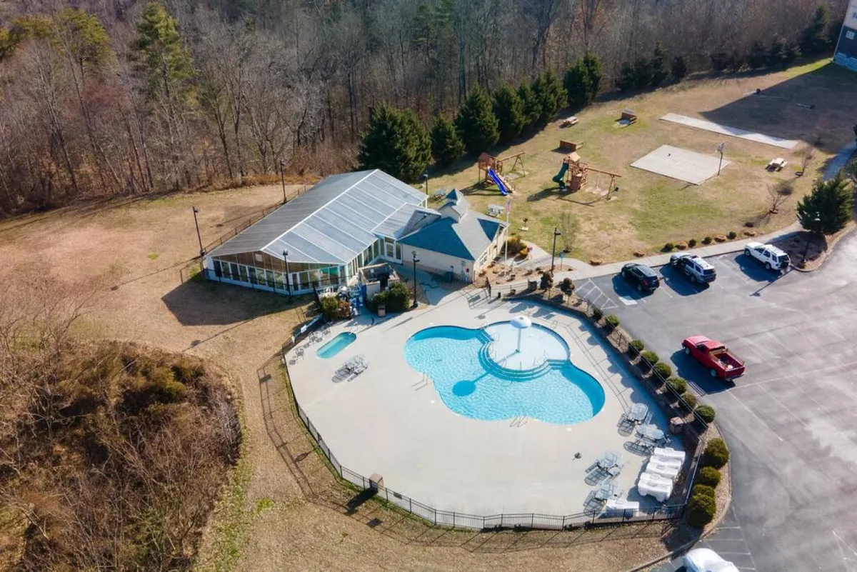 An inviting outdoor recreational area featuring a pool, adjacent playground, and surrounding amenities such as a parking lot and seating areas. Children are seen playing and families are gathered, conveying a lively, communal environment.