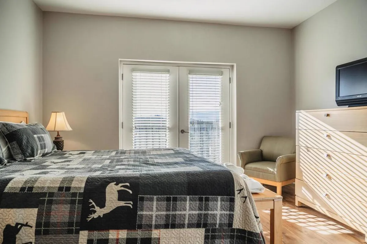 A serene bedroom with a bed covered in a patterned quilt, large windows with blinds that welcome sunrise views, an armchair, and a wall-mounted television, creating a space for relaxation, reading, or a cozy movie night.