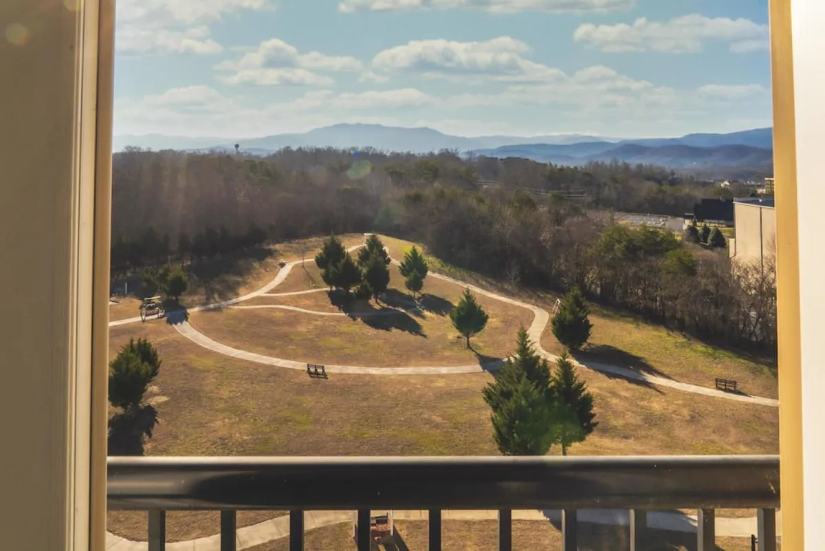 A tranquil resort view featuring curved walking paths shaded by green trees, benches placed along the trails, and mountains in the background. The overall mood is peaceful, ideal for relaxing walks or moments of connection in nature.