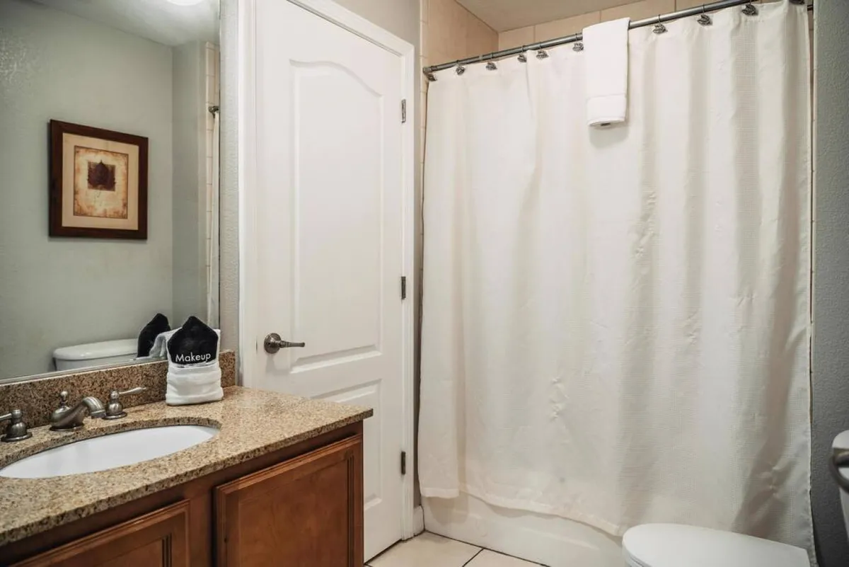 A modern bathroom with a granite countertop, white shower curtain, framed artwork, and a sink with a sleek faucet. The space feels tranquil and clean, ideal for refreshing morning routines or unwinding at night.