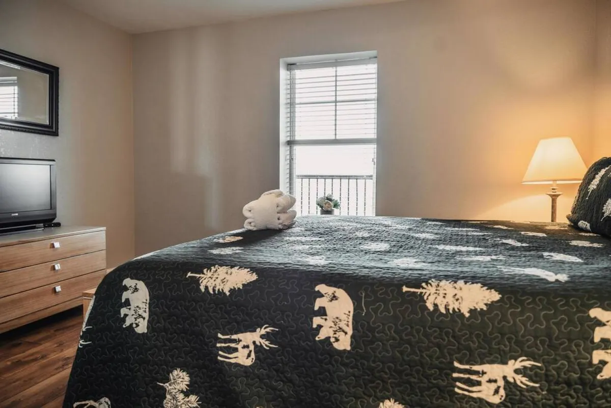 A cozy bedroom featuring a bed with a patterned quilt, nightstand with a lamp, a TV placed atop a dresser, and a window with blinds. The overall ambiance is inviting, perfect for unwinding and watching a favorite show before sleep.